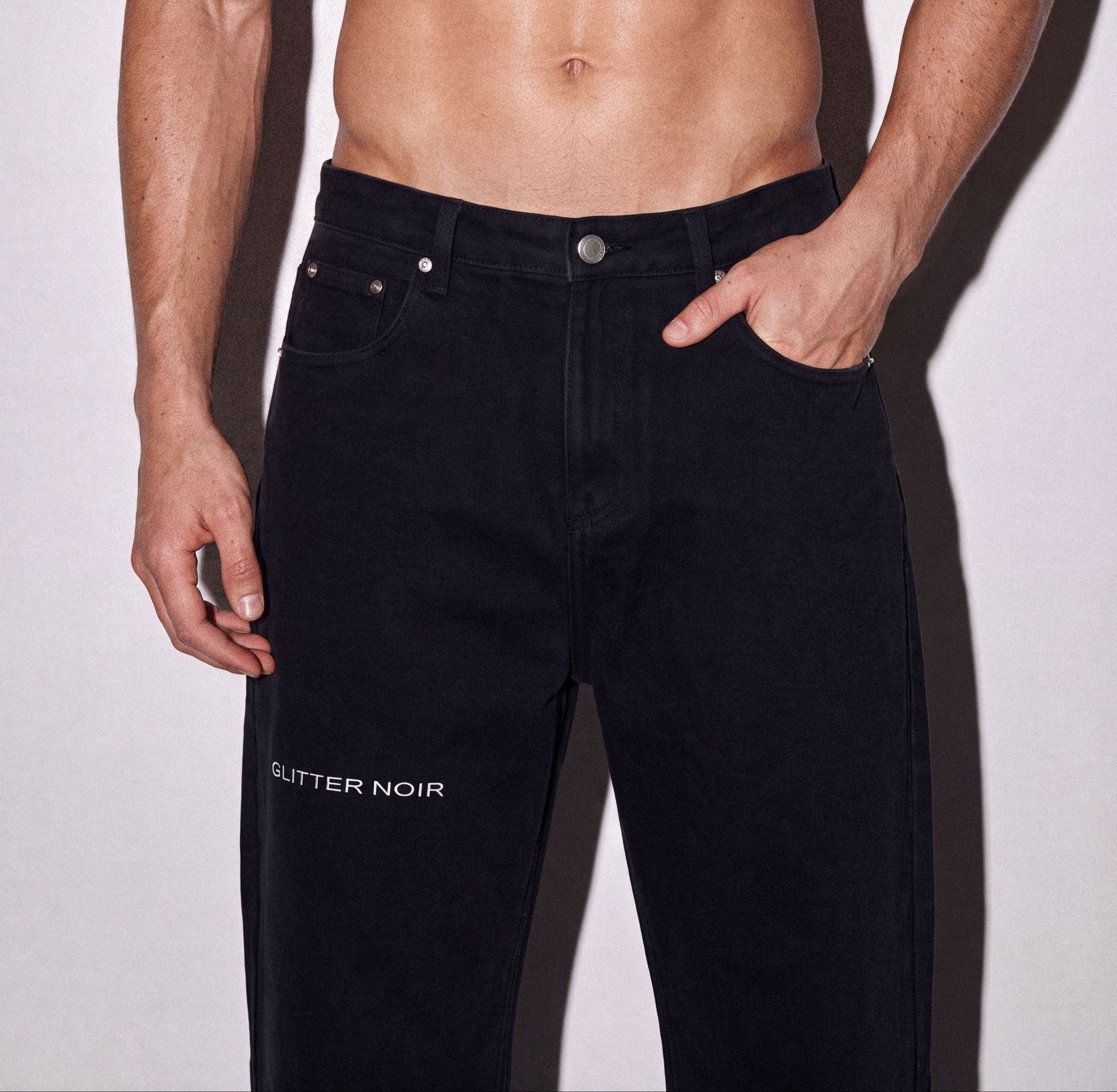 A shirtless muscular man wearing black denim jeans with 'GLITTER NOIR' printed in white on the left thigh, standing against a light gray background.